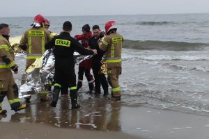 Plaża. Umundurowani ratownicy wodni i strażacy zakładają koce termiczne kobiecie i policjantowi.