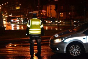 Pora nocna. Umundurowany policjant w żółtej kamizelce odblaskowej z napisem POLICJA stoi na ulicy, po której jadą samochody. Obok policjanta stoi oznakowany radiowóz