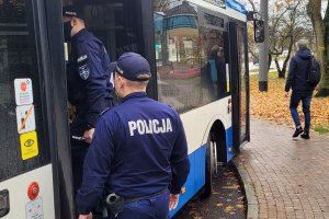 Pora jesienna. Umundurowani policjanci wchodzą do autobusu miejskiego. Za nimi w tle widoczny mężczyzna, który idzie po chodniku.