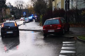 Fragment skrzyżowania o ruchu okrężnym, na którym stoi czerwony samochód częściowo na jezdni i na chodniku. Po rondzie jadą inne samochody. W tle w zatoczce autobusowej widać dwa oznakowane radiowozy.