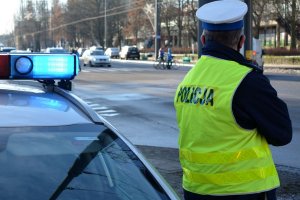 Zdjęcie archiwalne. Pora zimowa. Umundurowany policjant stoi przy radiowozie i obserwuje ruch pojazdów, pieszych i rowerzystów poruszających się ulicą.