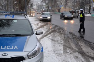 Zdjęcie archiwalne. Pora zimowa. Umundurowany policjant zatrzymuje do kontroli kierującego samochodem. Obok nich ulica, po której jadą inne pojazdy.