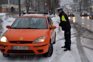 Zdjęcie archiwalne. Pora zimowa. Umundurowany policjant zatrzymał do kontroli kierującego samochodem. Obok nich ulica, po której jadą inne pojazdy.