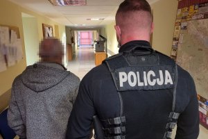 Policjant w stroju cywilnym z założoną kamizelką z napisem POLICJA prowadzi korytarzem zatrzymanego mężczyznę