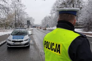 Pora zimowa, na drzewach śnieg. Umundurowany policjant stoi przy jezdni i obserwuje jadące pojazdy. Za nim na poboczu zaparkowany oznakowany radiowóz.