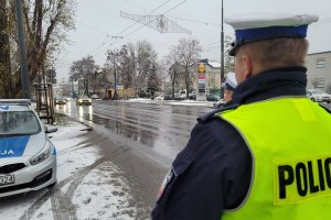 Pora zimowa, na drzewach śnieg. Umundurowani policjanci stoją przy jezdni i obserwują jadące pojazdy. Obok nich oznakowany radiowóz.