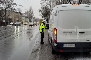 Umundurowany policjant stoi przy jezdni i rozmawia z kierującym białym busem. Obok nich ulica, po której jadą pojazdy.