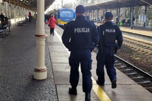 Peron dworca. Dwaj umundurowani policjanci idą peronem w stronę pociągu. Za nimi na peronach widoczne osoby