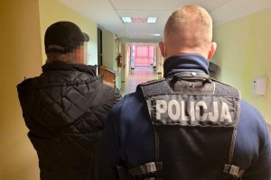 Policjant w stroju cywilnym i z założoną kamizelka taktyczną z napisem POLICJA prowadzi korytarzem zatrzymanego mężczyznę