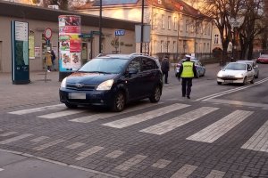 Przejście dla pieszych, na którym stoi samochód osobowy toyota, obok niego idzie umundurowany policjant. W tle idące chodnikiem osoby, jadące samochody i zaparkowany na chodniku oznakowany radiowóz.