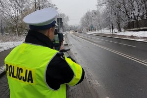 Pora zimowa. Umundurowany policjant stoi przy jezdni i za pomocą urządzenia trzymanego w rękach mierzy prędkość jadącego samochodu. Zdjęcie poglądowe.