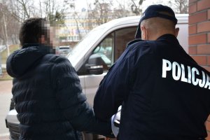 Umundurowany policjant prowadzi zatrzymanego mężczyznę. Za nimi bus.