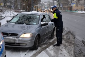 Pora zimowa. Umundurowany policjant stoi przy pojeździe i przeprowadza kontrolę drogową. Obok jezdnia, po której w tle jadą inne pojazdy. Zdjęcie archiwalne.