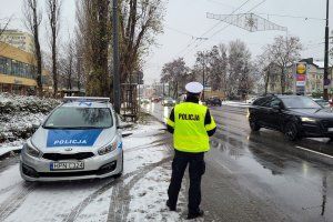 Pora zimowa, na ziemi leży śnieg. Umundurowany policjant stoi przy jezdni i obserwuje jadące ulicą samochody. Obok policjanta stoi zaparkowany w zatoczce oznakowany radiowóz.