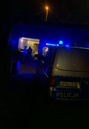 Pora nocna, teren zielony przy tunelu pod torami kolejowymi. Na pierwszym planie oznakowany radiowozów. W tle samochód i stojący przed nim umundurowany policjant i osoba w stroju cywilnym.