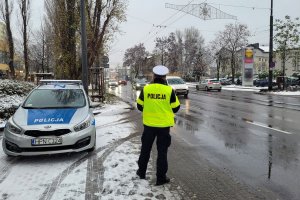 Pora zimowa. Umundurowany policjant stoi przy ulicy i obserwuje jadące ulicą samochody. Po lewej stronie od policjanta w zatoczce stoi oznakowany radiowóz.