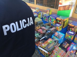 Fragment sylwetki umundurowanego policjanta, który na tyle kurtki ma napis POLICJA. Policjant stoi przy stoisku, na którym leżą opakowania z fajerwerkami.