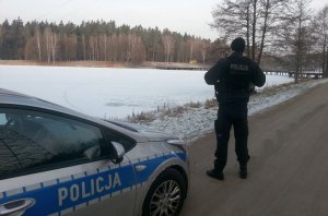 Umundurowany policjant stoi na drodze przy oznakowanym radiowozie i obserwuje widoczny za nim zamarznięty zbiornik wodny. W tle widoczny las.