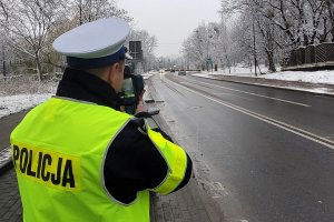 Pora zimowa. Umundurowany policjant stoi przy jezdni i za pomocą urządzenia trzymanego w rękach mierzy prędkość jadących ulicą pojazdów