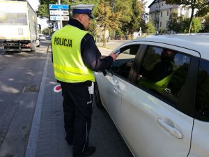 Umundurowany policjant stoi przy białym pojeździe i trzyma w ręku urządzenie do badania stanu trzeźwości.