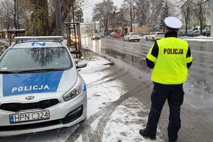 Pora zimowa. Umundurowany policjant stoi przy ulicy obok zaparkowanego oznakowanego radiowozu i obserwuje samochody jadące ulicą.