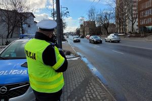 Umundurowany policjant stoi przy jedni i trzymając w ręce urządzenie mierzy prędkość samochodów jadących ulicą. Za policjantem z lewej strony znajduje się oznakowany radiowóz