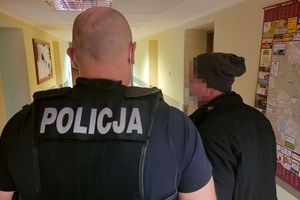 Policjant w stroju cywilnym i z założoną kamizelką taktyczną z napisem POLICJA prowadzi korytarzem zatrzymanego mężczyznę.