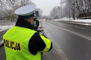 Pora zimowa. Umundurowany policjant stoi przy ulicy, trzyma w ręku urządzenie i mierzy prędkość jadących ulicą pojazdów.