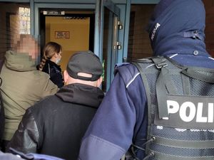 Umundurowani policjanci wprowadzają do budynku dwóch mężczyzn ubranych w stroje cywilne.