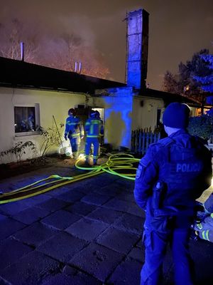 Umundurowany policjant stoi przed budynkiem i zabezpiecza miejsce działań strażaków. Za policjantem widoczne sylwetki trzech strażaków, którzy wchodzą do pomieszczenia budynku.