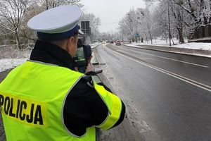 Pora zimowa. Umundurowany policjant stoi przy jezdni i za pomocą trzymanego w ręku urządzenia mierzy prędkość pojazdu.