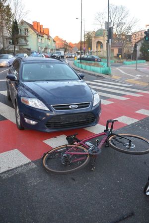 Pora dzienna. Na zdjęciu ulica, na której znajduje się przejazd dla rowerów i przejście dla pieszych. Na przejściu dla pieszych i przejeździe dla rowerów stoi samochód, a przed nim leży rower. Za samochodem widoczny fragment oznakowanego radiowozu i inne samochody