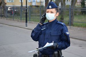 Umundurowana policjantka stoi przy ulicy i rozmawia przez telefon, który trzyma w jednej dłoni. W drugiej dłoni trzyma akta.