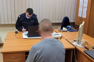 Pomieszczenie służbowe. Umundurowany policjant siedząc za biurkiem wypisuje mandat mężczyźnie, który siedzi na krześle.