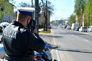 Umundurowany policjant stoi przy jezdni i trzymając w ręku urządzenie mierzy nim prędkość jadących pojazdów - zdjęcie archiwalne.