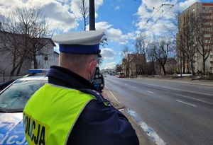 Umundurowany policjant stoi przy ulicy, trzyma w rękach urządzenie i mierzy prędkość jadących ulicą pojazdów