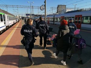 Peron dworca kolejowego. Umundurowani policjanci razem z grupą osób w strojach cywilnych idą po peronie.