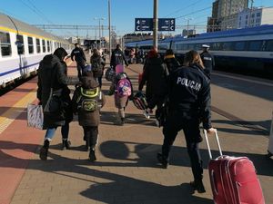 Peron dworca kolejowego. Umundurowani policjanci razem z grupą osób w strojach cywilnych idą po peronie. Policjanci niosą i ciągną bagaże.