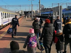 Peron dworca kolejowego. Umundurowani policjanci razem z grupą osób w strojach cywilnych idą po peronie. Policjanci niosą bagaże.