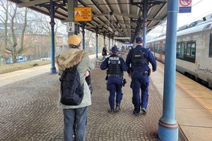 Peron dworca kolejowego. Umundurowani policjanci idą wzdłuż peronu obok innych osób stojących na peronie.