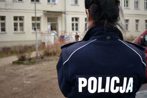 Umundurowana policjantka stoi przy budynku i obserwuje osoby stojące przed budynkiem