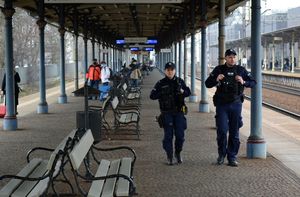 Peron dworca kolejowego. Umundurowani policjanci idą wzdłuż peronu obok innych osób stojących na peronie.