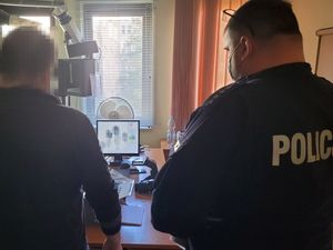 Pomieszczenie służbowe. Umundurowany policjant stoi obok zatrzymanego mężczyzny.