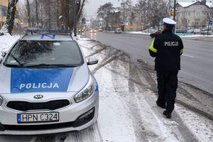 Pora zimowa. Umundurowany policjant stoi przy ulicy i obserwuje zachowania kierujących pojazdami jadących ulicą. Z lewej strony w zatoczce stoi oznakowany radiowóz. - Zdjęcie archiwalne