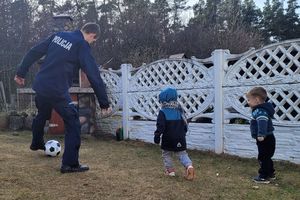 Ogrodzone podwórko, na którym przebywa umundurowany policjant i dwoje dzieci, z którymi gra w piłkę