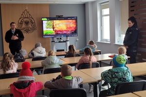 Pomieszczenie remizy strażackiej. Policjantka i strażak rozmawiają z grupą dzieci