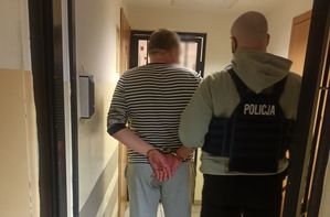 Policjant w stroju cywilnym i z założoną kamizelką z napisem POLICJA wprowadza do pomieszczenia dla osób zatrzymanych mężczyznę