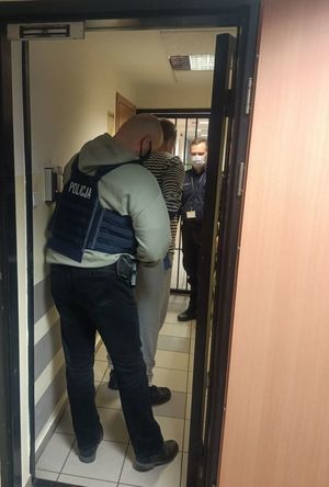 Policjant w stroju cywilnym i z założoną kamizelką z napisem POLICJA wprowadza do pomieszczenia dla osób zatrzymanych mężczyznę