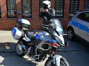 Umundurowany policjant stoi przy motocyklu i przygotowuje się do jazdy. Obok widać fragment oznakowanego radiowozu.