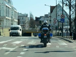 Umundurowany policjant na oznakowanym motocyklu jedzie ulicą. Obok widoczne inne pojazdy i budynki